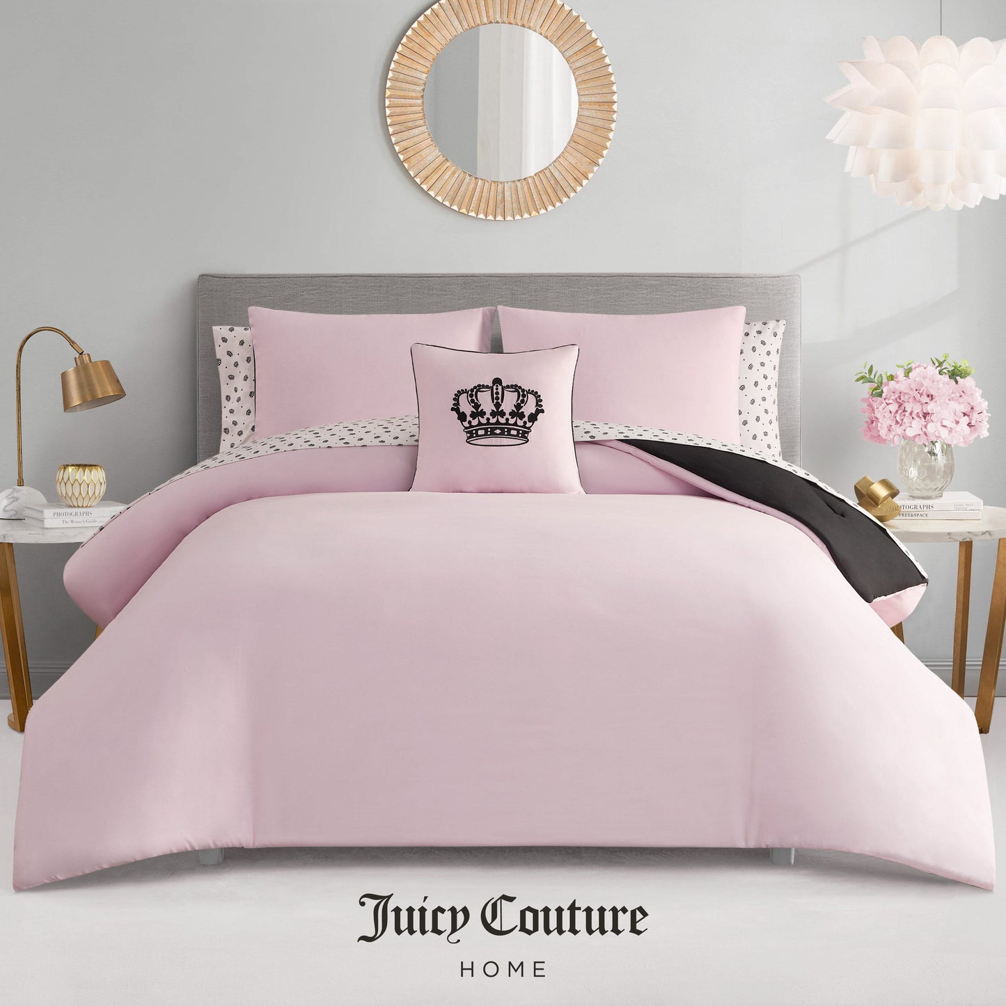 A neatly made bed features the Crown Sparkle Solid Reversible Bed in a Bag Bedding Set, pink with polka dot pillowcases and a black crown decorative pillow. The glam room includes a round mirror, flowers, and Juicy Couture Home branding below.