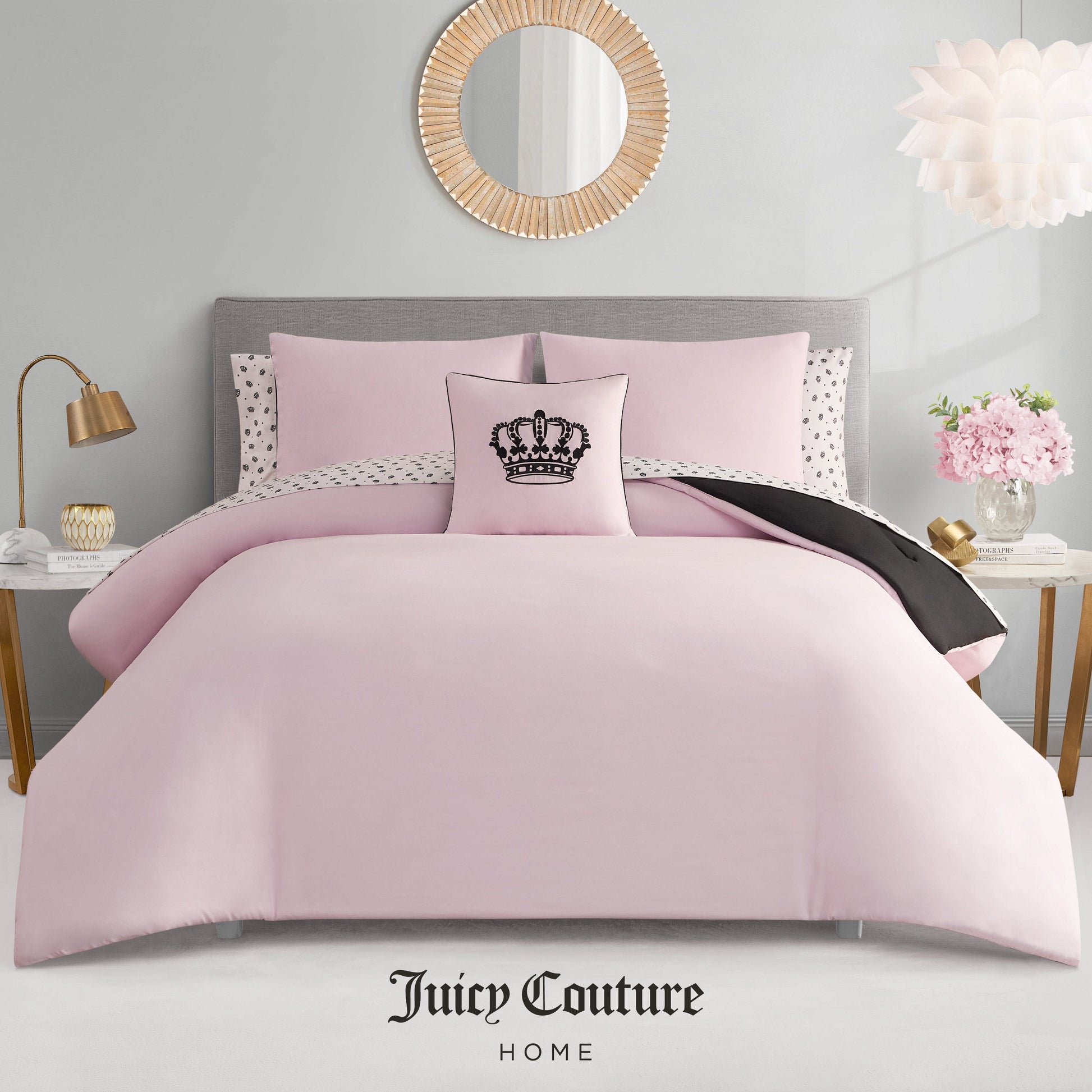 A neatly made bed features the Crown Sparkle Solid Reversible Bed in a Bag Bedding Set, pink with polka dot pillowcases and a black crown decorative pillow. The glam room includes a round mirror, flowers, and Juicy Couture Home branding below.