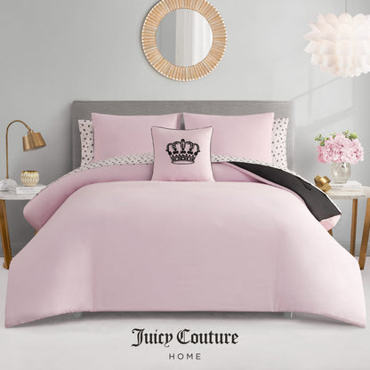 A neatly made bed features the Crown Sparkle Solid Reversible Bed in a Bag Bedding Set, pink with polka dot pillowcases and a black crown decorative pillow. The glam room includes a round mirror, flowers, and Juicy Couture Home branding below.