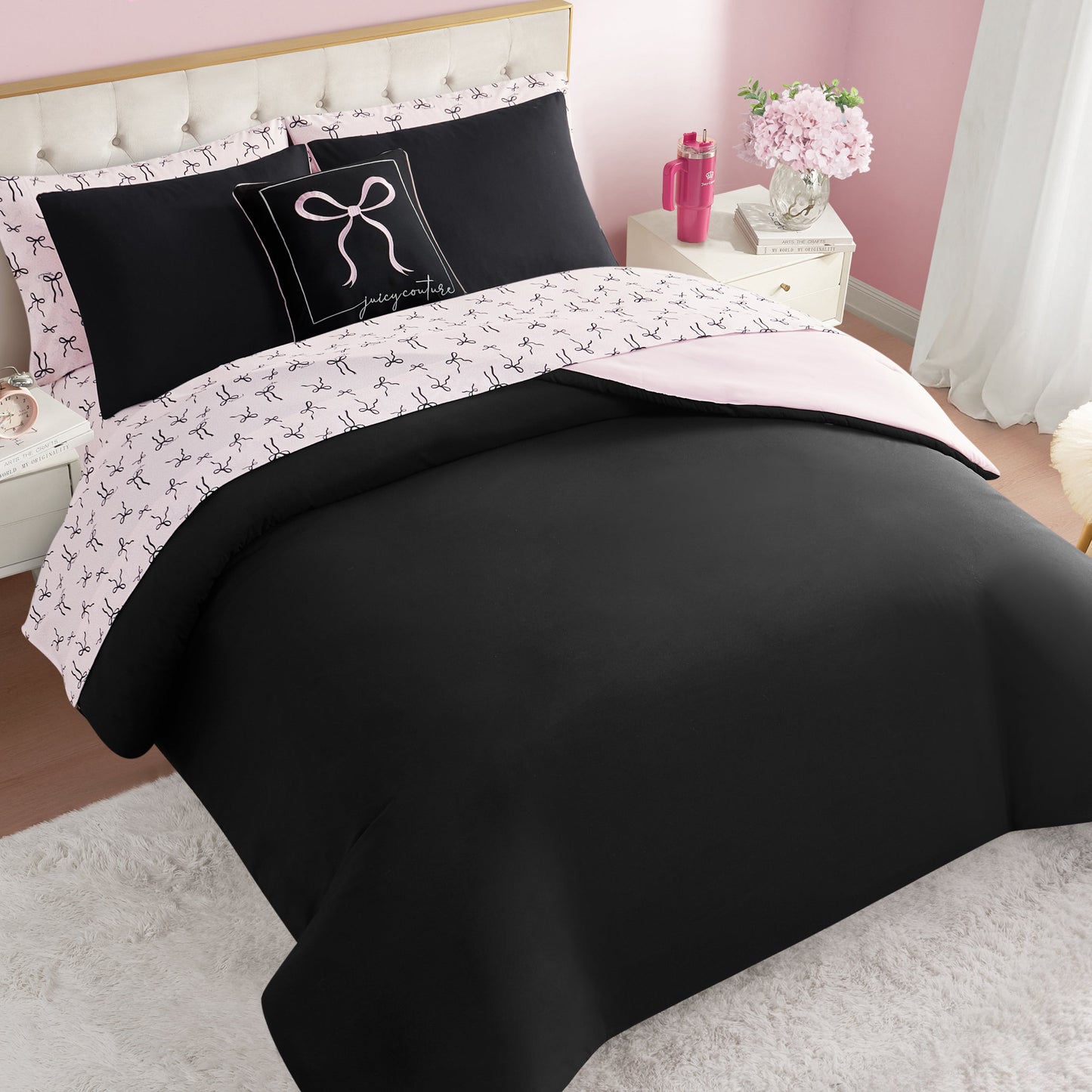 A neatly made bed with the Bows Solid Reversible Bed in a Bag Bedding Set in black and pale pink, featuring bow-patterned sheets and a decorative pillow. The room has pink walls, white nightstands, and flowers in a vase.