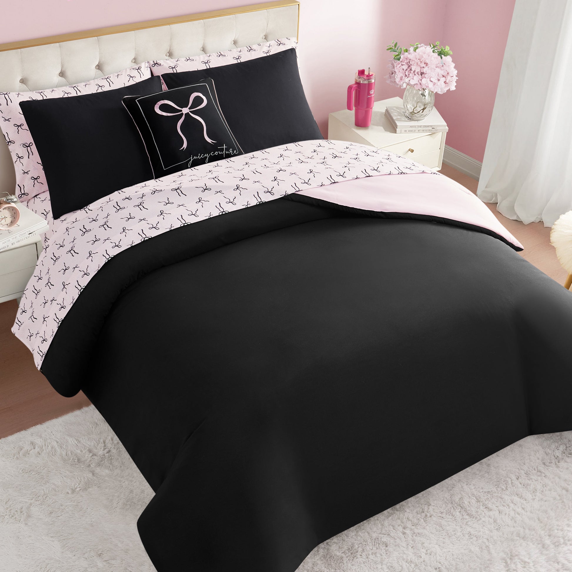 A neatly made bed with the Bows Solid Reversible Bed in a Bag Bedding Set in black and pale pink, featuring bow-patterned sheets and a decorative pillow. The room has pink walls, white nightstands, and flowers in a vase.