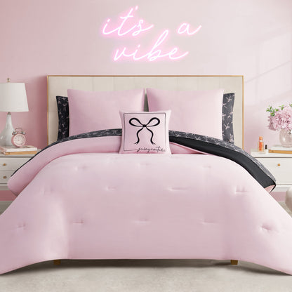 A neatly made bed with the Bows Solid Reversible Bed in a Bag Bedding Set and a decorative pillow featuring a black bow and text. The room has light pink walls, a white nightstand with lamp, flowers, and a neon "its a vibe" sign above the bed.