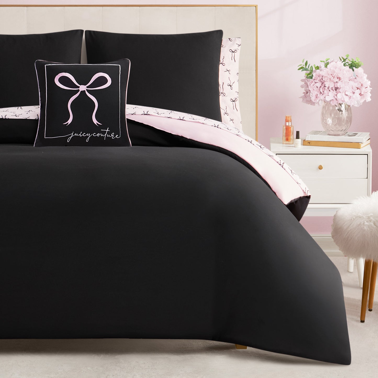 A neatly made bed features the Bows Solid Reversible Bed in a Bag Bedding Set with black bedding, a matching pillow with a pink bow and Juicy Couture text, light pink sheet trim, and a white nightstand holding flowers and a bottle.