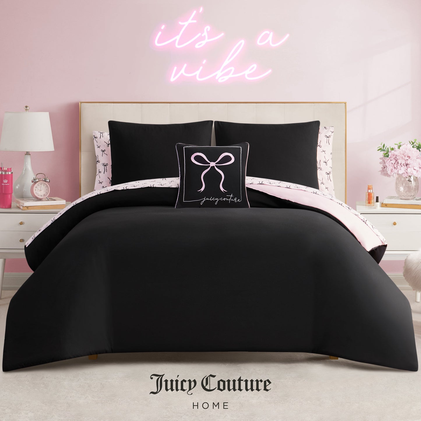 A stylish bedroom featuring a beige tufted headboard, the Juicy Couture Bows Solid Reversible Bed in a Bag Bedding Set in black with light pink accents, a decorative pillow, and a neon pink "it’s a vibe" sign on the light pink wall.