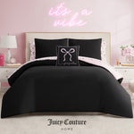 A stylish bedroom featuring a beige tufted headboard, the Juicy Couture Bows Solid Reversible Bed in a Bag Bedding Set in black with light pink accents, a decorative pillow, and a neon pink "it’s a vibe" sign on the light pink wall.