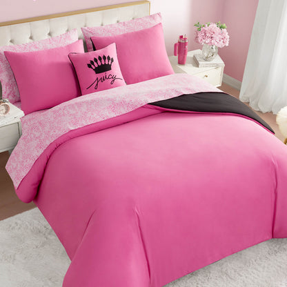 A neatly made bed with the In The Wild Solid Reversible Bed in a Bag set, featuring a reversible comforter, pink and white sheets, matching pillows—including an accent pillow with a black crown and “juicy”—plus pink decor and white furniture.