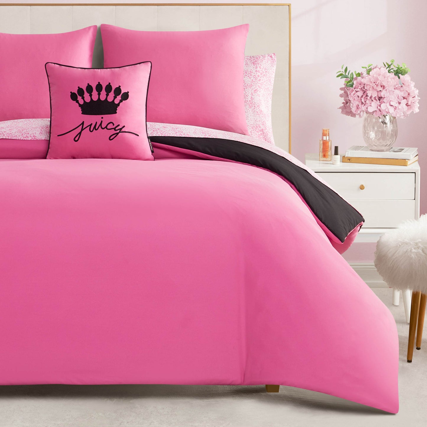 A neatly made bed features bright pink bedding from the In The Wild Solid Reversible Bed in a Bag set, accented with a black and pink crown pillow and reversible comforter. A white nightstand with pink flowers, books, and perfume sits beside it.