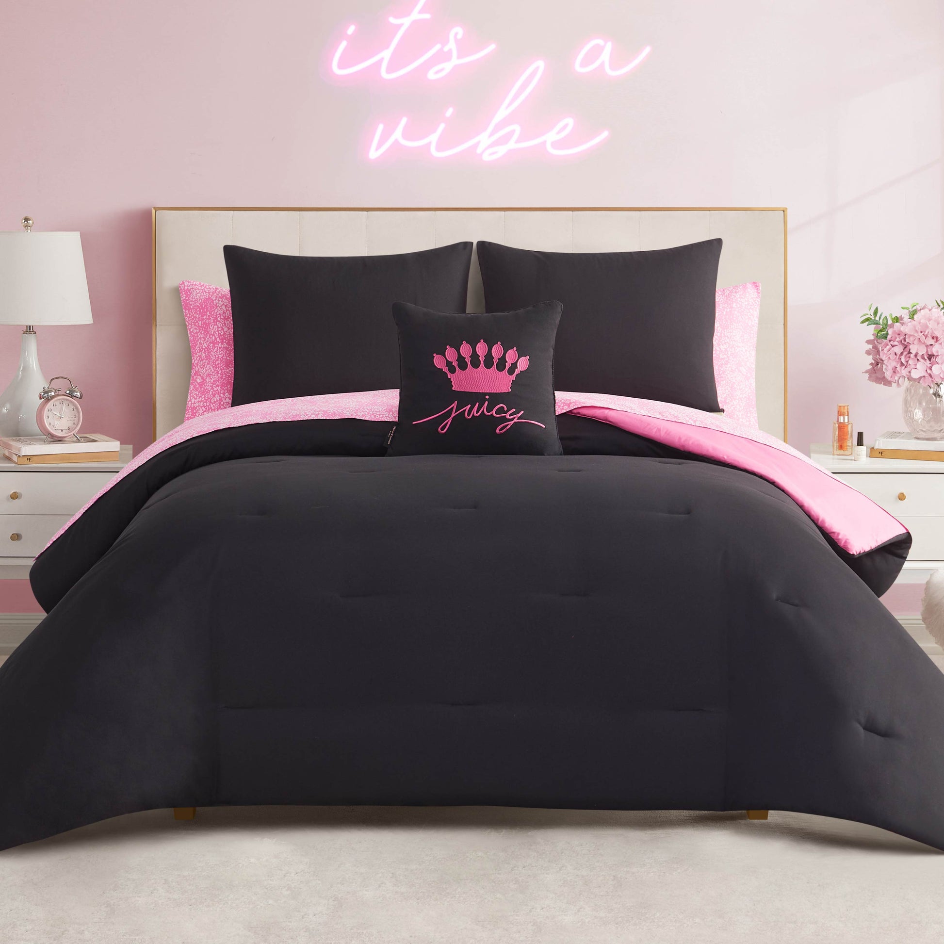 A neatly made bed features the In The Wild Solid Reversible Bed in a Bag Bedding Set in black and pink, with a decorative "Juicy" pillow. The room has a pink wall, neon sign, and white nightstands with lamps.