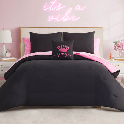 A neatly made bed features the In The Wild Solid Reversible Bed in a Bag Bedding Set in black and pink, with a decorative "Juicy" pillow. The room has a pink wall, neon sign, and white nightstands with lamps.