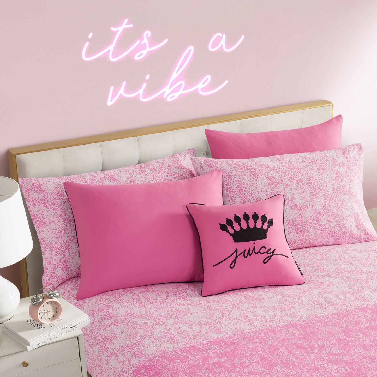 A pink-themed bedroom features the In The Wild Solid Reversible Bed in a Bag Bedding Set with pink patterned sheets, solid pink pillows, and a small pillow with a black crown and "Juicy" text. Neon lights above the bed spell "its a vibe" on the wall.