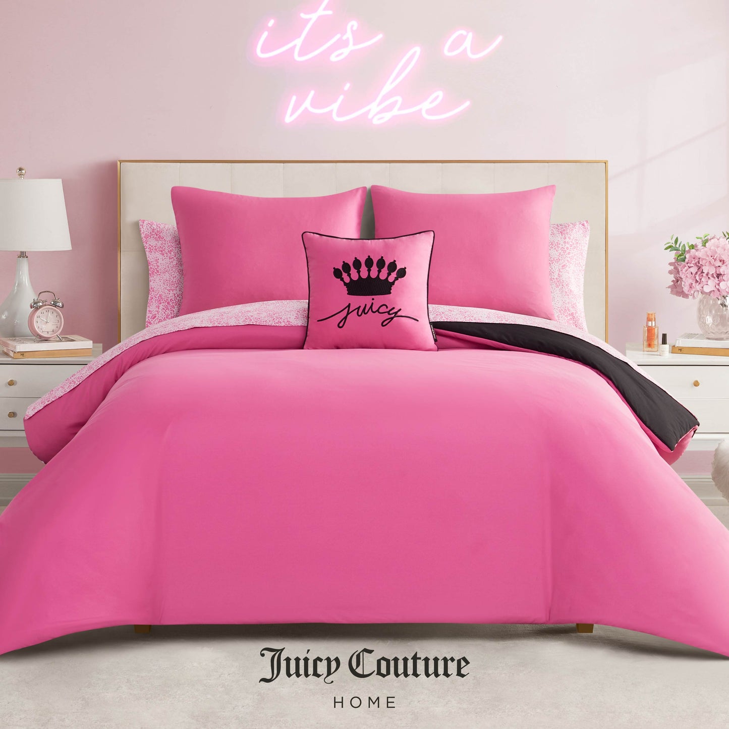 A stylish bedroom features the Juicy Couture Home In The Wild Solid Reversible Bed in a Bag Bedding Set with bright pink bedding and a "Juicy" crown pillow. A pink neon "it’s a vibe" sign glows on the wall behind. Juicy Couture Home logo at bottom.