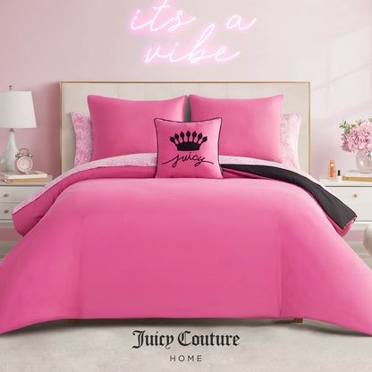 A stylish bedroom features the Juicy Couture Home In The Wild Solid Reversible Bed in a Bag Bedding Set with bright pink bedding and a "Juicy" crown pillow. A pink neon "it’s a vibe" sign glows on the wall behind. Juicy Couture Home logo at bottom.