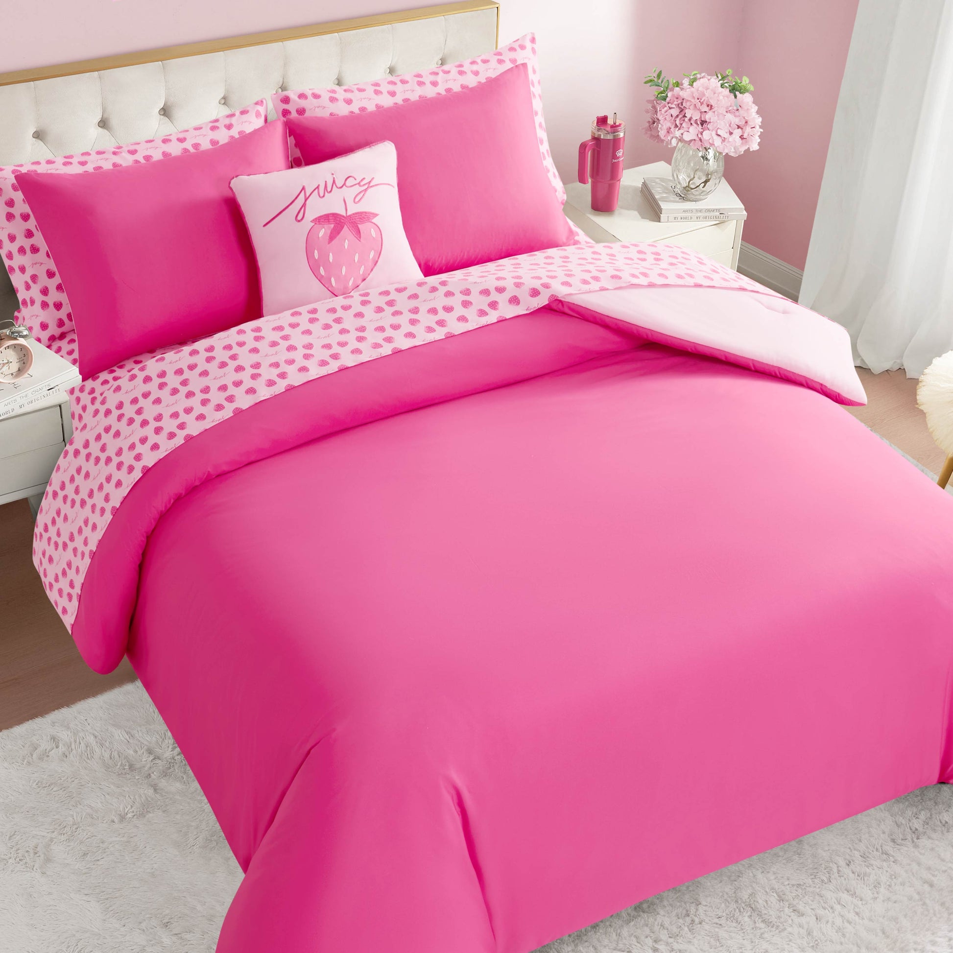A neatly made bed features the Summer Strawberries Solid Reversible Bed in a Bag Bedding Set with a bright pink comforter, heart-patterned sheets, and strawberry-accented pillows. The room has pink walls and white furniture.