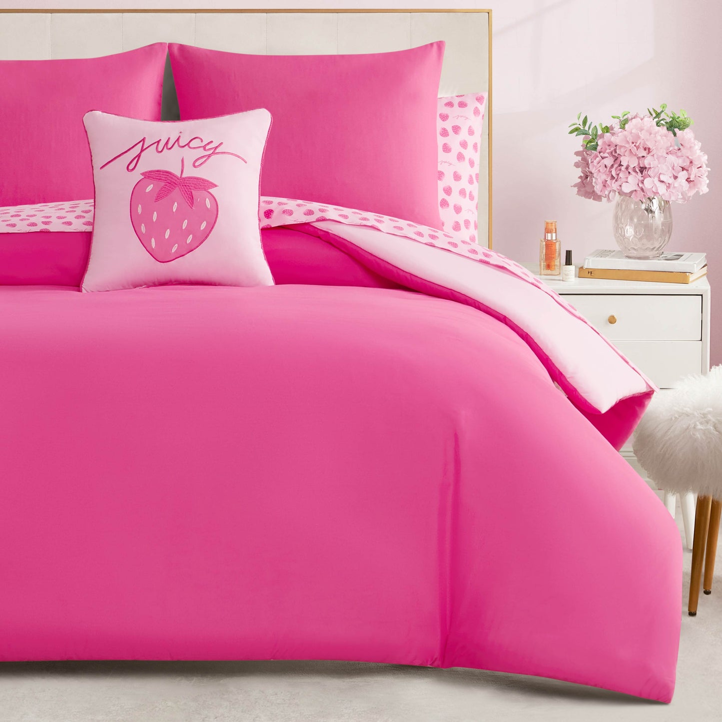 A Summer Strawberries Solid Reversible Bed in a Bag Bedding Set with bright pink strawberry bedding and a themed pillow sits beside a white nightstand with pink flowers and a fluffy white stool nearby.