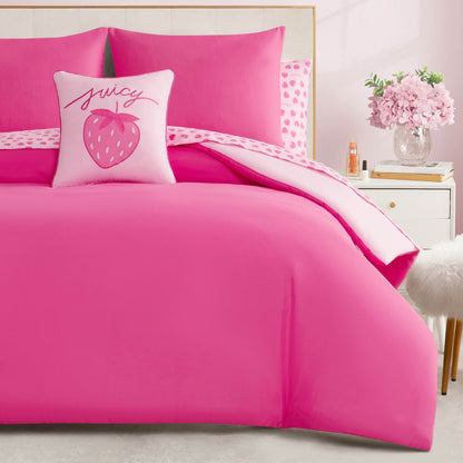 A Summer Strawberries Solid Reversible Bed in a Bag Bedding Set with bright pink strawberry bedding and a themed pillow sits beside a white nightstand with pink flowers and a fluffy white stool nearby.