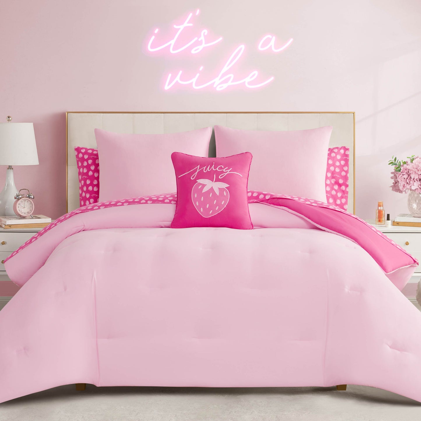 A neatly made bed features the Summer Strawberries Solid Reversible Bed in a Bag Bedding Set and a strawberry throw pillow with "juicy," set against a pink wall with a neon "it's a vibe" sign, flanked by nightstands with lamps and decor.