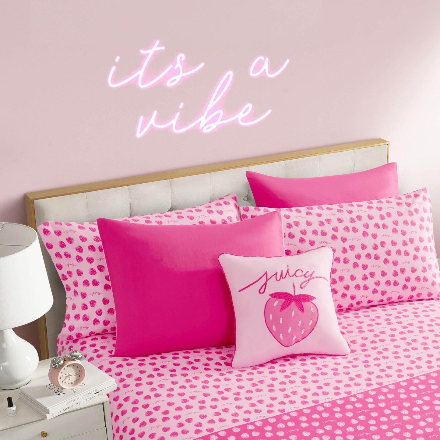The Summer Strawberries Solid Reversible Bed in a Bag Bedding Set features pink spotted strawberry bedding, bright and light pink pillows, plus a decorative pillow with a strawberry and "juicy," paired with a neon "it’s a vibe" sign above the bed.