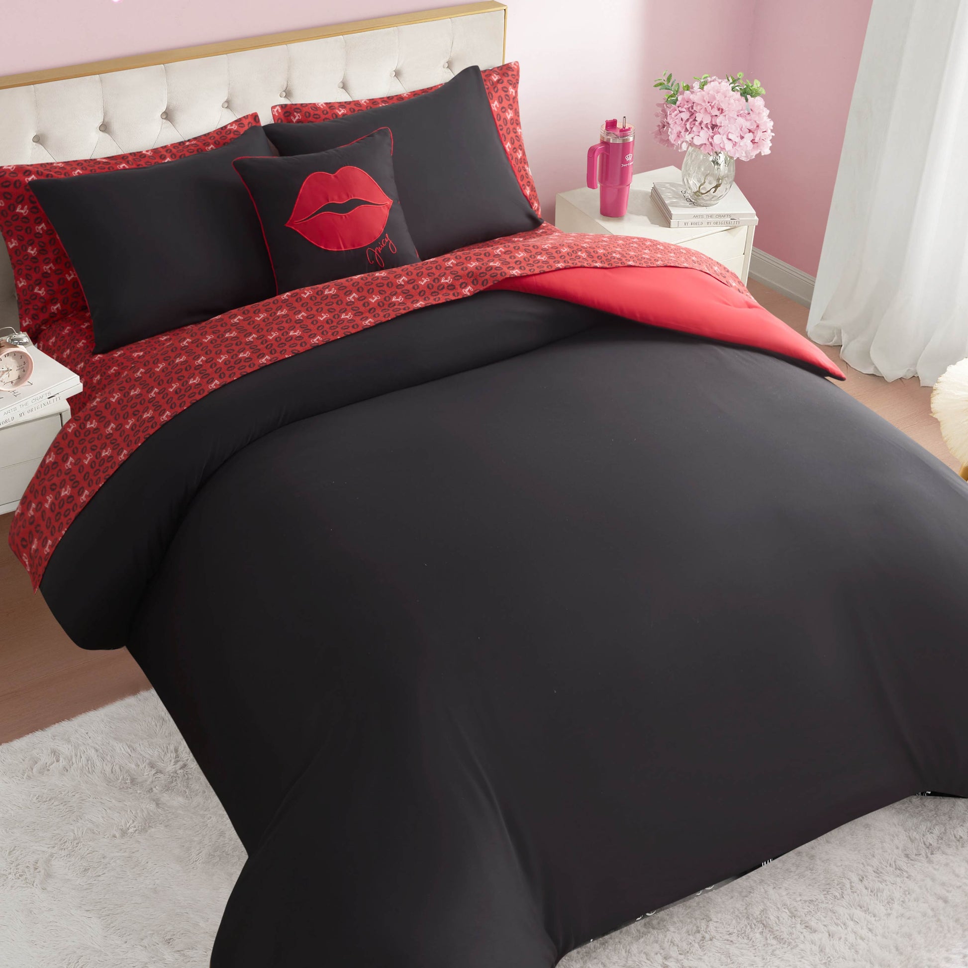 A neatly made bed features the Cheetah Rebel Lips Solid Reversible Bed in a Bag Bedding Set, with pillows displaying a red lip design, set against a tufted headboard in a pink room with flowers, a nightstand, and white curtains.
