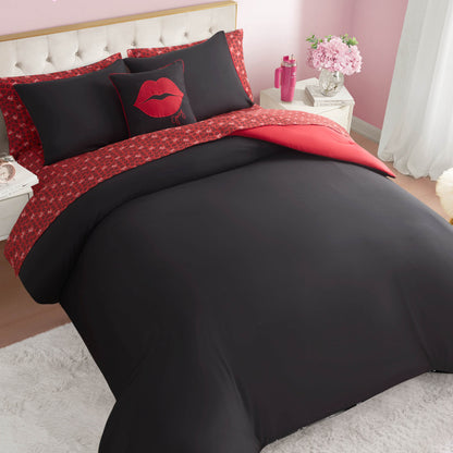 A neatly made bed features the Cheetah Rebel Lips Solid Reversible Bed in a Bag Bedding Set, with pillows displaying a red lip design, set against a tufted headboard in a pink room with flowers, a nightstand, and white curtains.