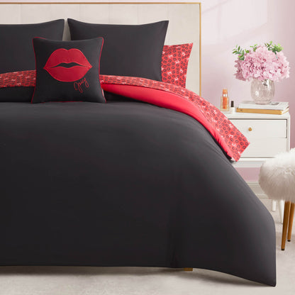 A Juicy Couture Cheetah Rebel Lips Solid Reversible Bed in a Bag Bedding Set with black and red bedding and a red lips pillow is displayed in a bright room with a white nightstand, pink flowers, and a fluffy white stool.