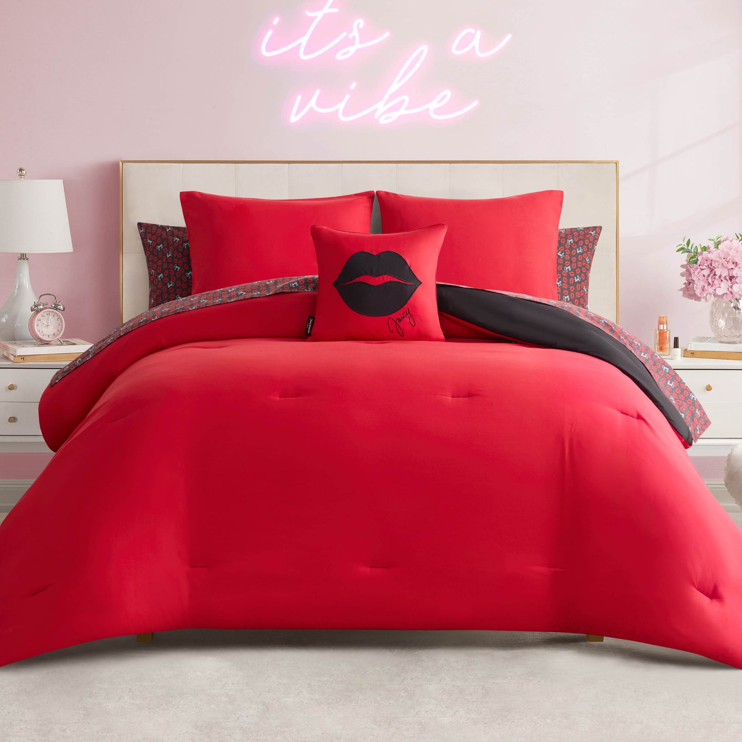A Juicy Couture Cheetah Rebel Lips Solid Reversible Bed in a Bag Bedding Set neatly made with vibrant red bedding and pillows, including a black lips pillow. Pink walls, a bedside lamp, and a neon “it’s a vibe” sign complete the room.