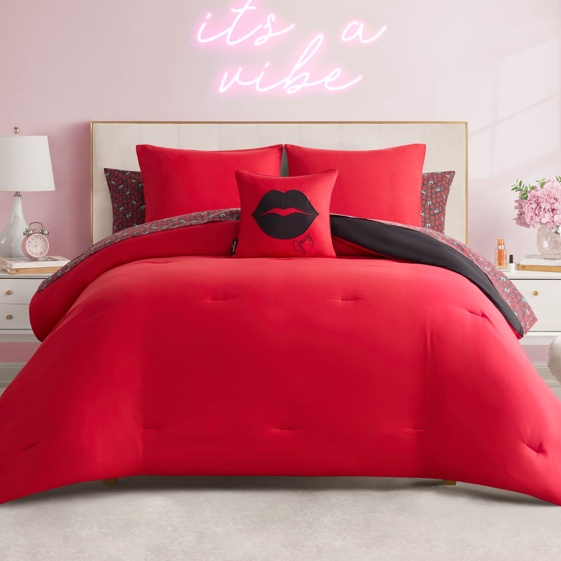 A Juicy Couture Cheetah Rebel Lips Solid Reversible Bed in a Bag Bedding Set neatly made with vibrant red bedding and pillows, including a black lips pillow. Pink walls, a bedside lamp, and a neon “it’s a vibe” sign complete the room.