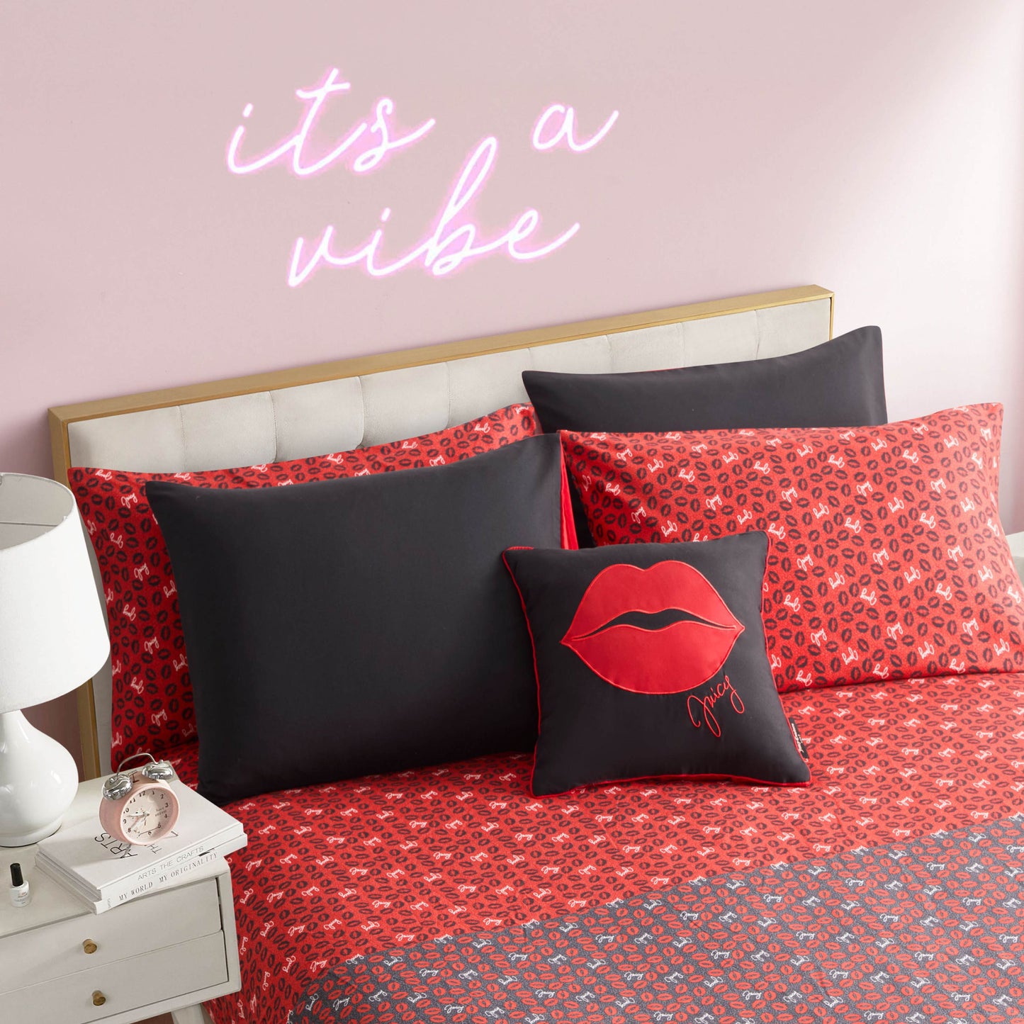 A Cheetah Rebel Lips Solid Reversible Bed in a Bag Bedding Set with red patterns and pillows, including a black pillow with red lips, sits under a pink wall and neon "its a vibe" sign. A nightstand nearby holds a lamp and clock.