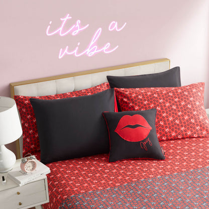 A Cheetah Rebel Lips Solid Reversible Bed in a Bag Bedding Set with red patterns and pillows, including a black pillow with red lips, sits under a pink wall and neon "its a vibe" sign. A nightstand nearby holds a lamp and clock.
