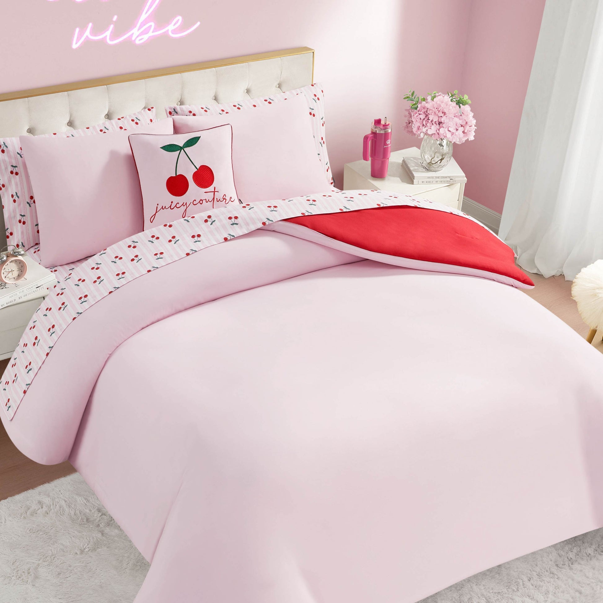 A neatly made bed features the Cherry Script Solid Reversible Bed in a Bag Bedding Set and a cherry-themed pillow with a red heart accent. Pink walls, a white nightstand topped with pink flowers, and white curtains finish this charming room.