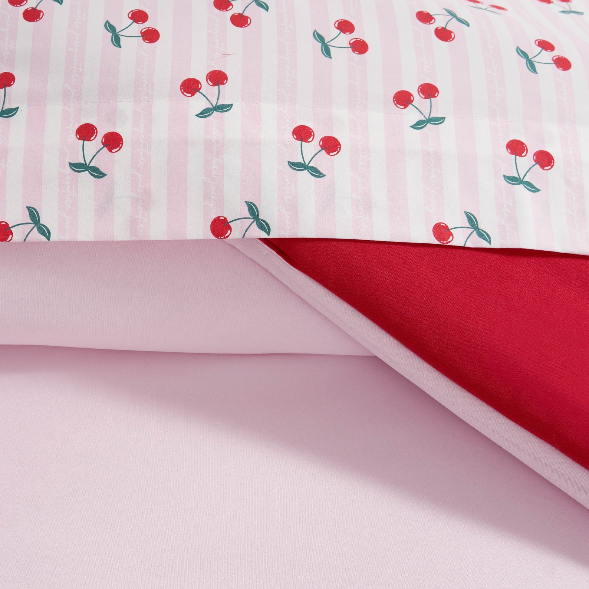 Close-up of the Cherry Script Solid Reversible Bed in a Bag Bedding Set: features a pink and white striped top sheet with red cherries, a plain red sheet underneath, and a light pink bottom sheet—pairs perfectly with your favorite comforter.