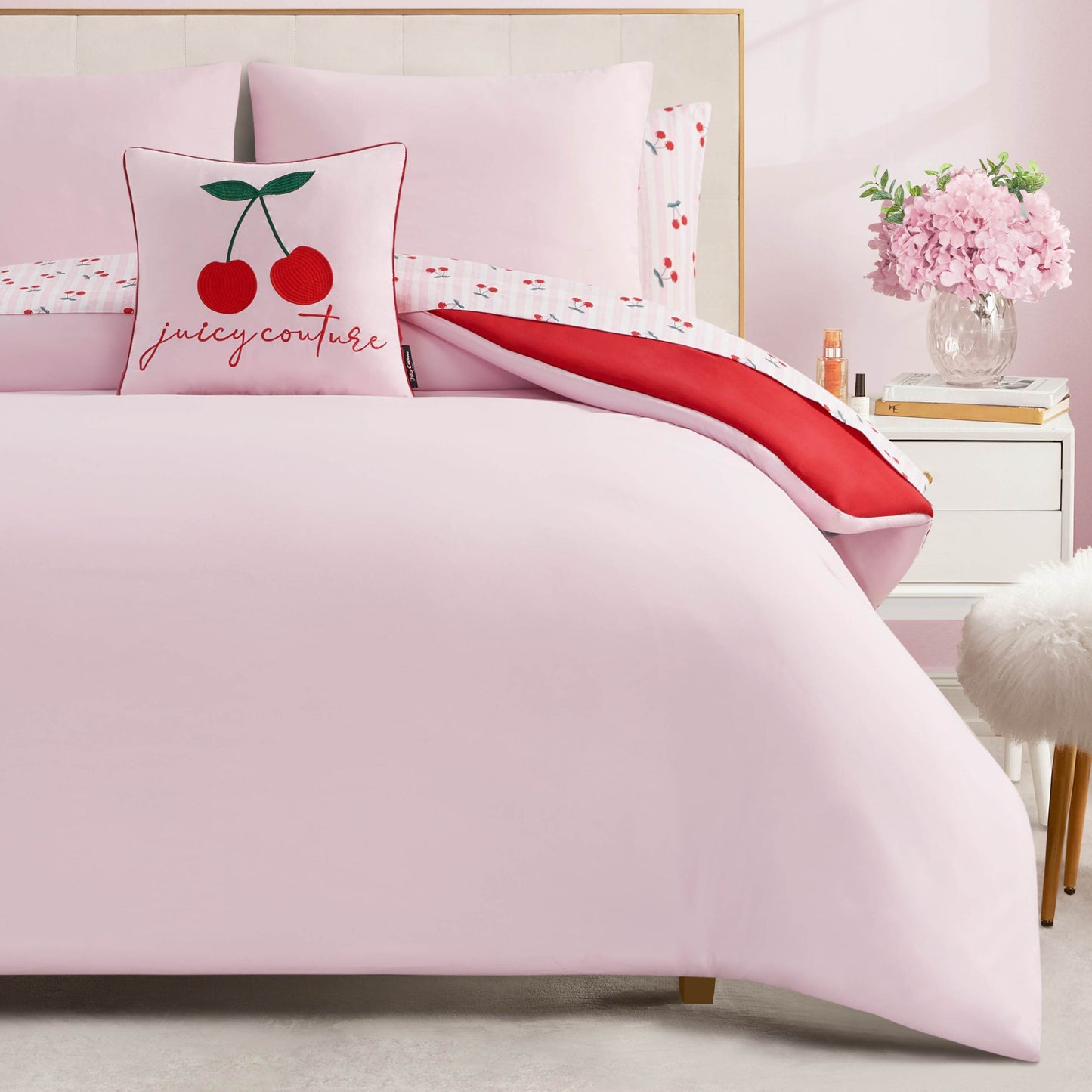 A neatly made bed with the Cherry Script Solid Reversible Bed in a Bag Bedding Set and a Juicy Couture cherry pillow. The room includes a nightstand with pink flowers, books, and a fluffy white stool for added charm and comfort.