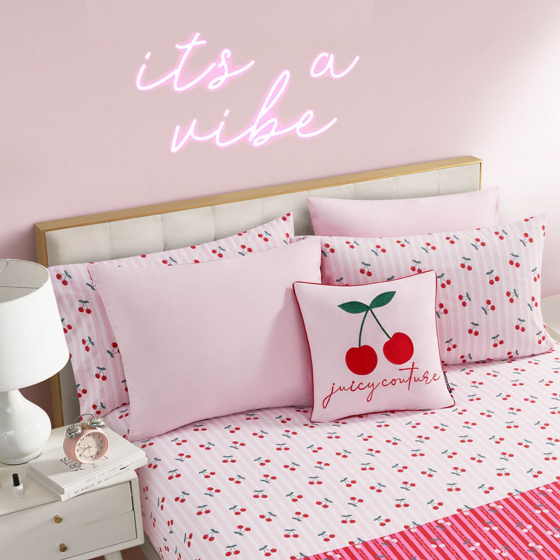 A neatly made bed with the Cherry Script Solid Reversible Bed in a Bag Bedding Set and pink pillows, plus a decorative cushion with cherries and "juicy couture." Above, a neon sign reads "it's a vibe," tying together this Juicy Couture comforter look.
