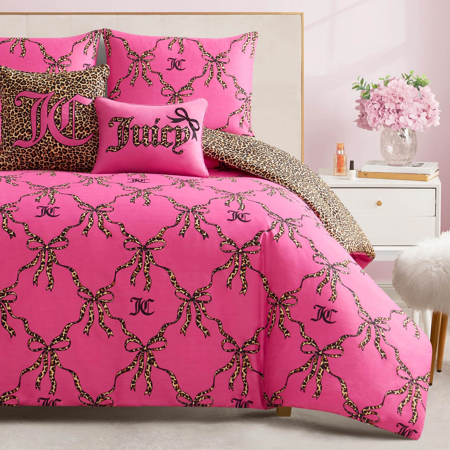 A bed with the Cheetah Bow Trellis Reversible Comforter Set in pink, featuring leopard-print bows and JC logos, plus two decorative pillows—one says Juicy, the other has a cheetah print—beside a white nightstand and a fluffy stool.
