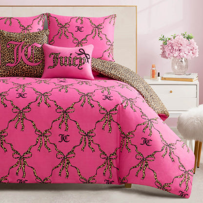 A bed with the Cheetah Bow Trellis Reversible Comforter Set in pink, featuring leopard-print bows and JC logos, plus two decorative pillows—one says Juicy, the other has a cheetah print—beside a white nightstand and a fluffy stool.