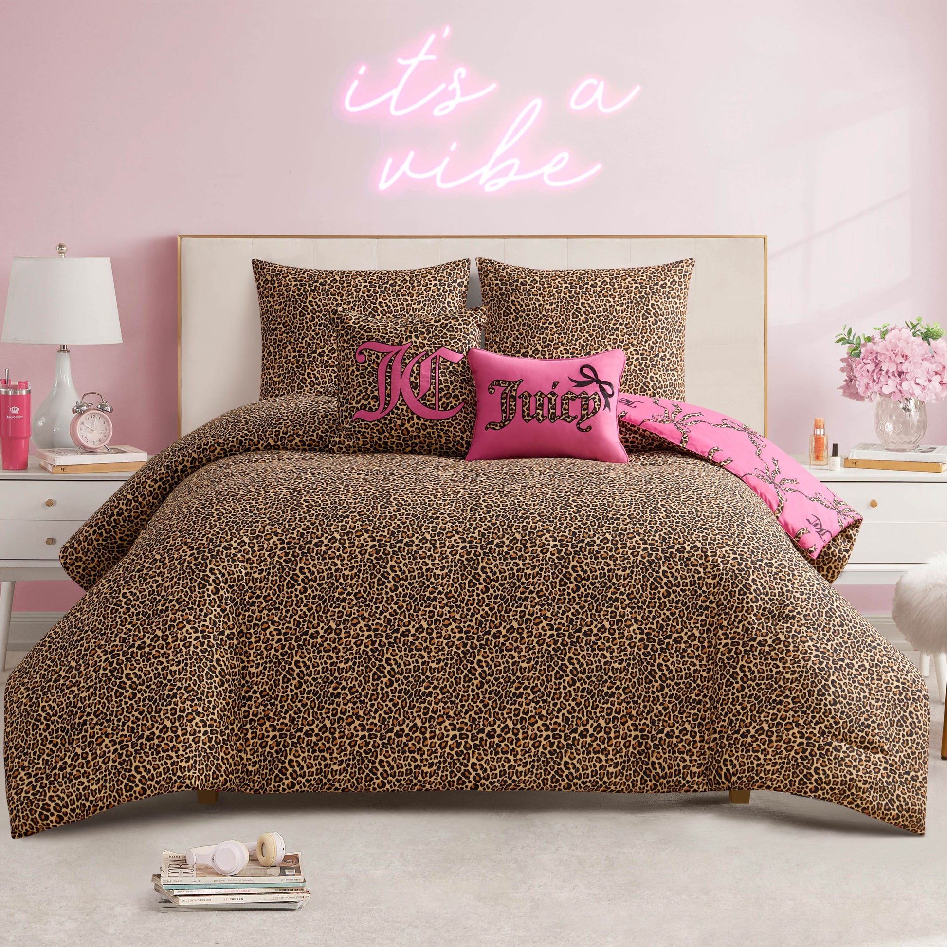 A bedroom featuring the Cheetah Bow Trellis Reversible Comforter Set, pink accent pillows, and an "it's a vibe" neon sign on a pink wall. White nightstands display lamps, flowers, and decor for a bold, fashion-inspired style.