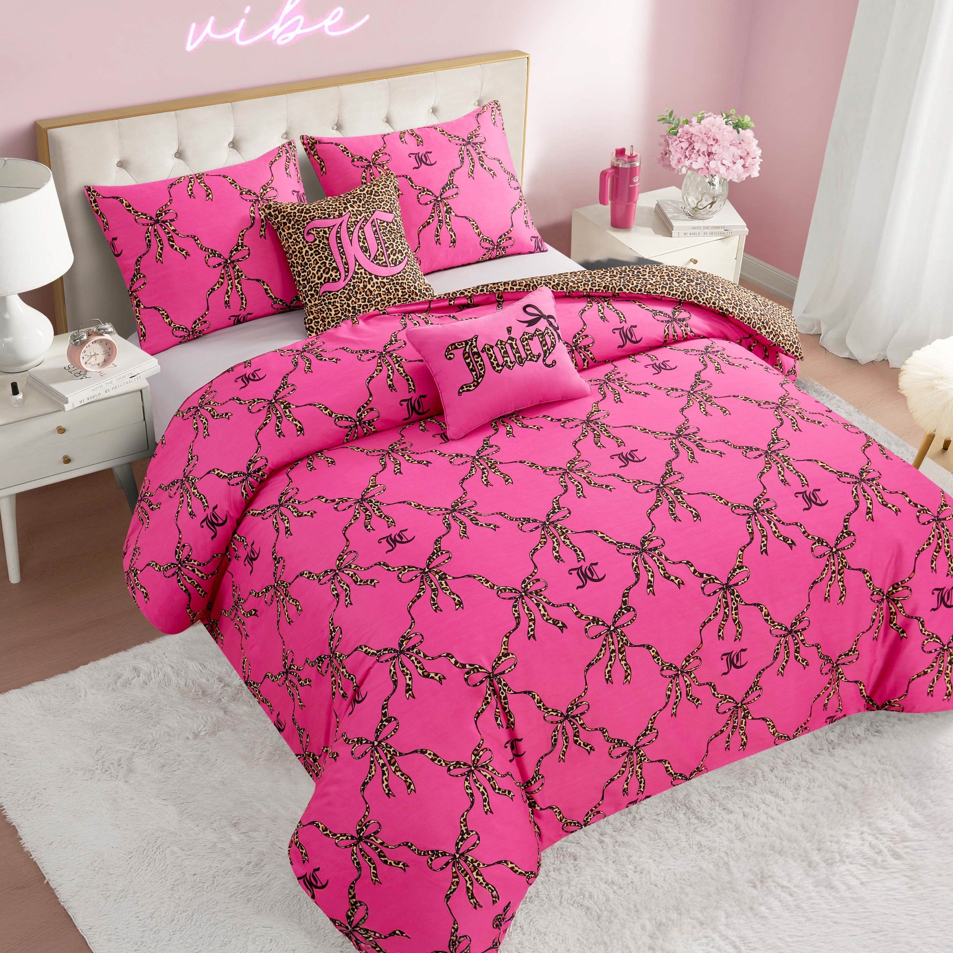 A pink bedroom features a bed with the Cheetah Bow Trellis Reversible Comforter Set and pillows adorned with cheetah-print chains, JC and Juicy logos. The room has white furniture, pink walls, and a "vibe" neon sign for a fashion-inspired look.