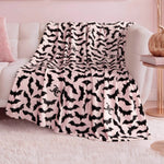 A Halloween Plush Throw Blanket 50" x 60" in soft pink and black with flying bats and a stylized “SC” is draped over a white couch, adding playful, cozy Halloween decor to the pastel-themed living room.