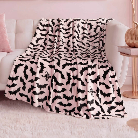 A Halloween Plush Throw Blanket 50" x 60" in soft pink and black with flying bats and a stylized “SC” is draped over a white couch, adding playful, cozy Halloween decor to the pastel-themed living room.