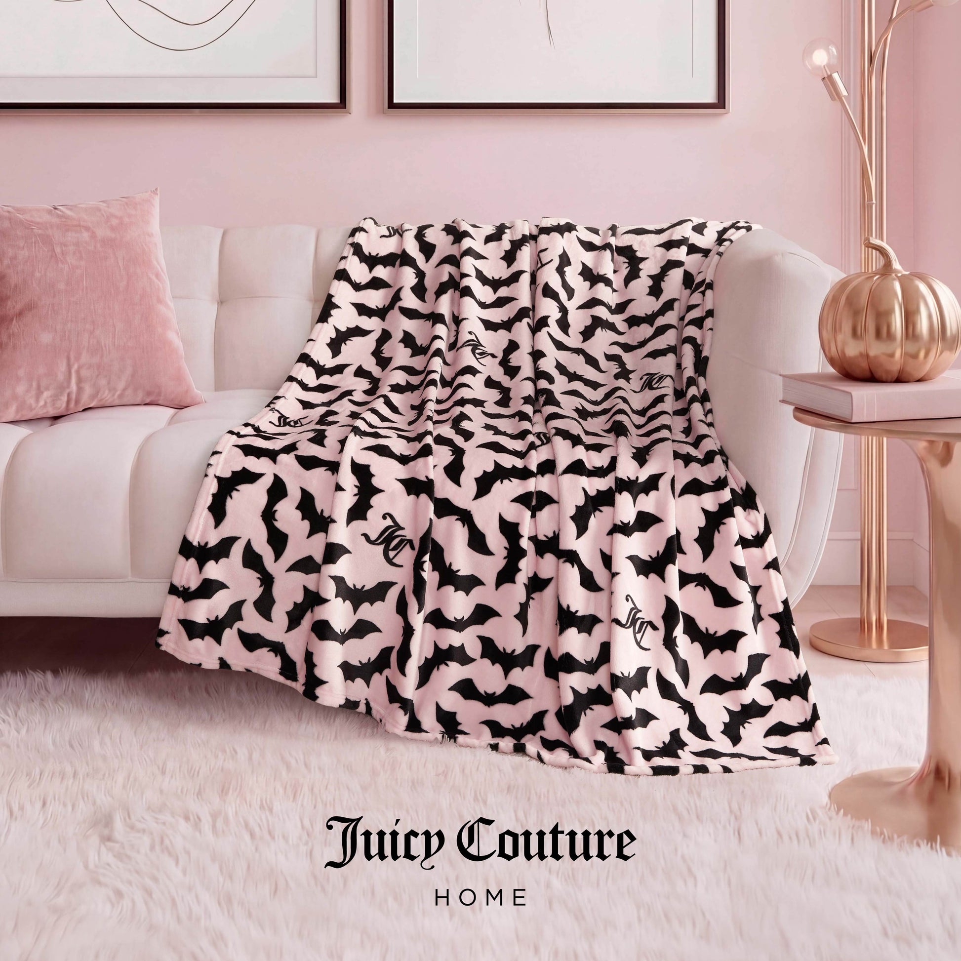 A Halloween Plush Throw Blanket 50" x 60" with a black bat pattern is draped over a white sofa in a pink room, adding playful Halloween decor. A cozy pink pillow and plush throw complete the inviting setup.