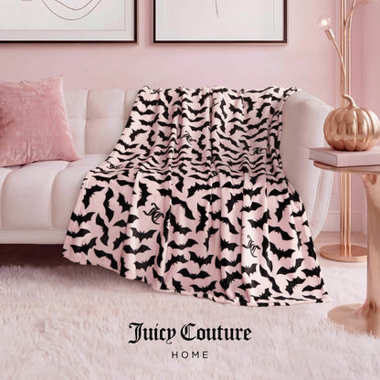 A Halloween Plush Throw Blanket 50" x 60" with a black bat pattern is draped over a white sofa in a pink room, adding playful Halloween decor. A cozy pink pillow and plush throw complete the inviting setup.