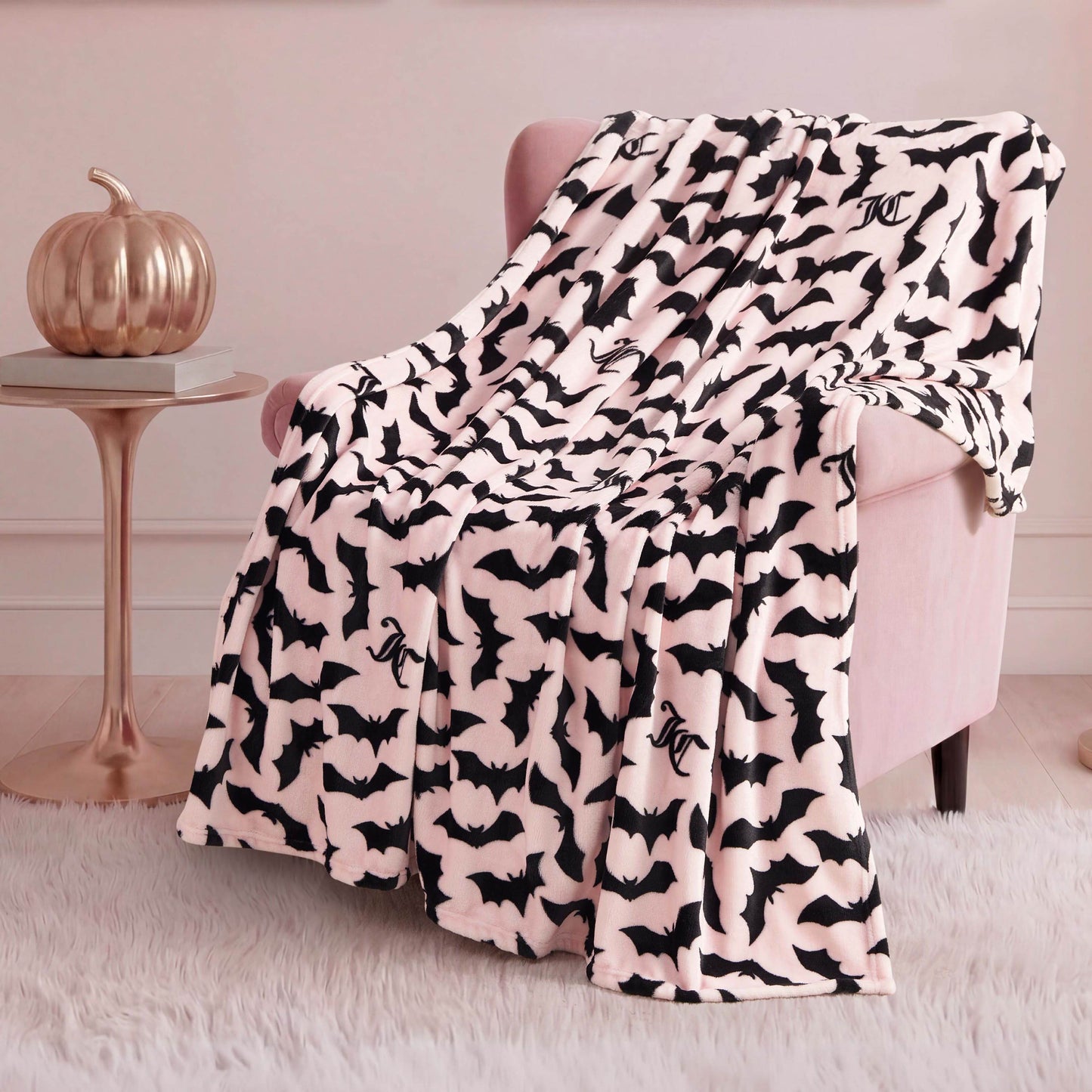 A soft, pale pink armchair is draped with the Halloween Plush Throw Blanket 50" x 60" featuring black bats, atop a white furry rug. A metallic pumpkin adds festive Halloween decor to the small side table in the background.