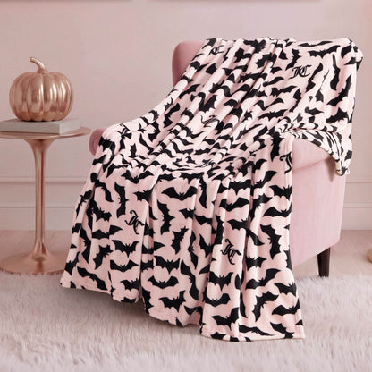 A soft, pale pink armchair is draped with the Halloween Plush Throw Blanket 50" x 60" featuring black bats, atop a white furry rug. A metallic pumpkin adds festive Halloween decor to the small side table in the background.