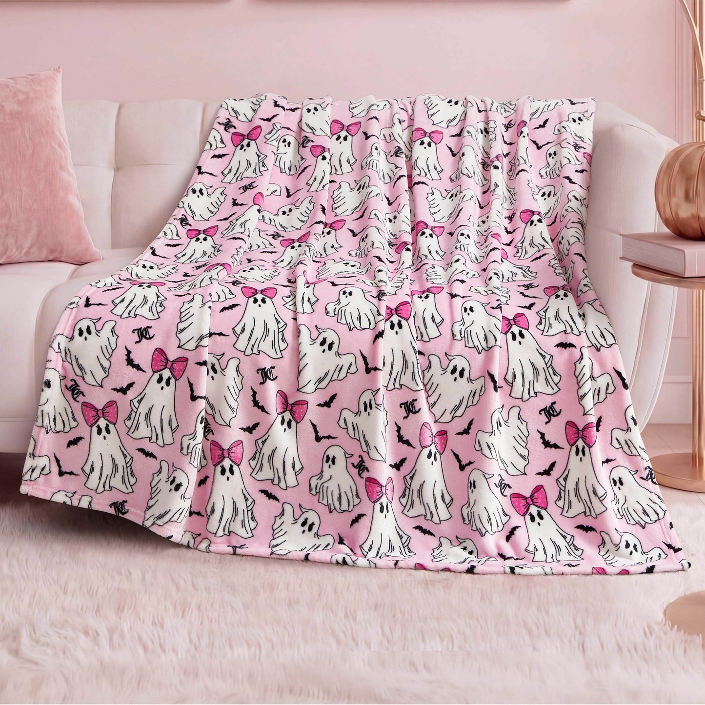 The Halloween Plush Throw Blanket 50" x 60", featuring cute white ghosts—some with pink bows—is draped over a white sofa in a pastel-themed room with pink decor and a fluffy rug, making it perfect for chic Halloween home decor.