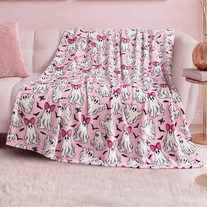 The Halloween Plush Throw Blanket 50" x 60", featuring cute white ghosts—some with pink bows—is draped over a white sofa in a pastel-themed room with pink decor and a fluffy rug, making it perfect for chic Halloween home decor.