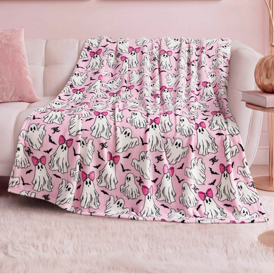 The Halloween Plush Throw Blanket 50" x 60", featuring cute white ghosts—some with pink bows—is draped over a white sofa in a pastel-themed room with pink decor and a fluffy rug, making it perfect for chic Halloween home decor.