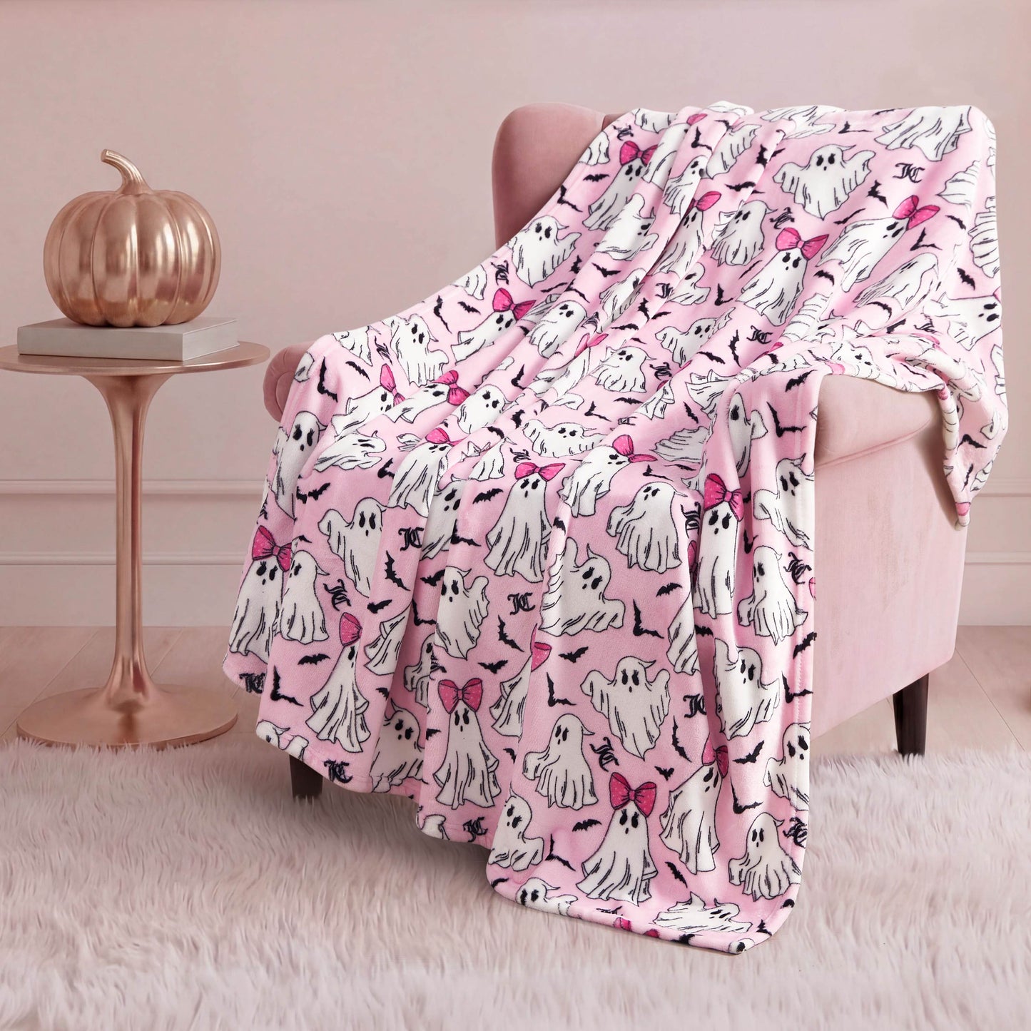 A pink armchair is draped with the Halloween Plush Throw Blanket 50" x 60", featuring cartoon ghosts, some with pink bows. A metallic pumpkin on a round side table and a white rug add a cozy, festive Halloween touch to the room.