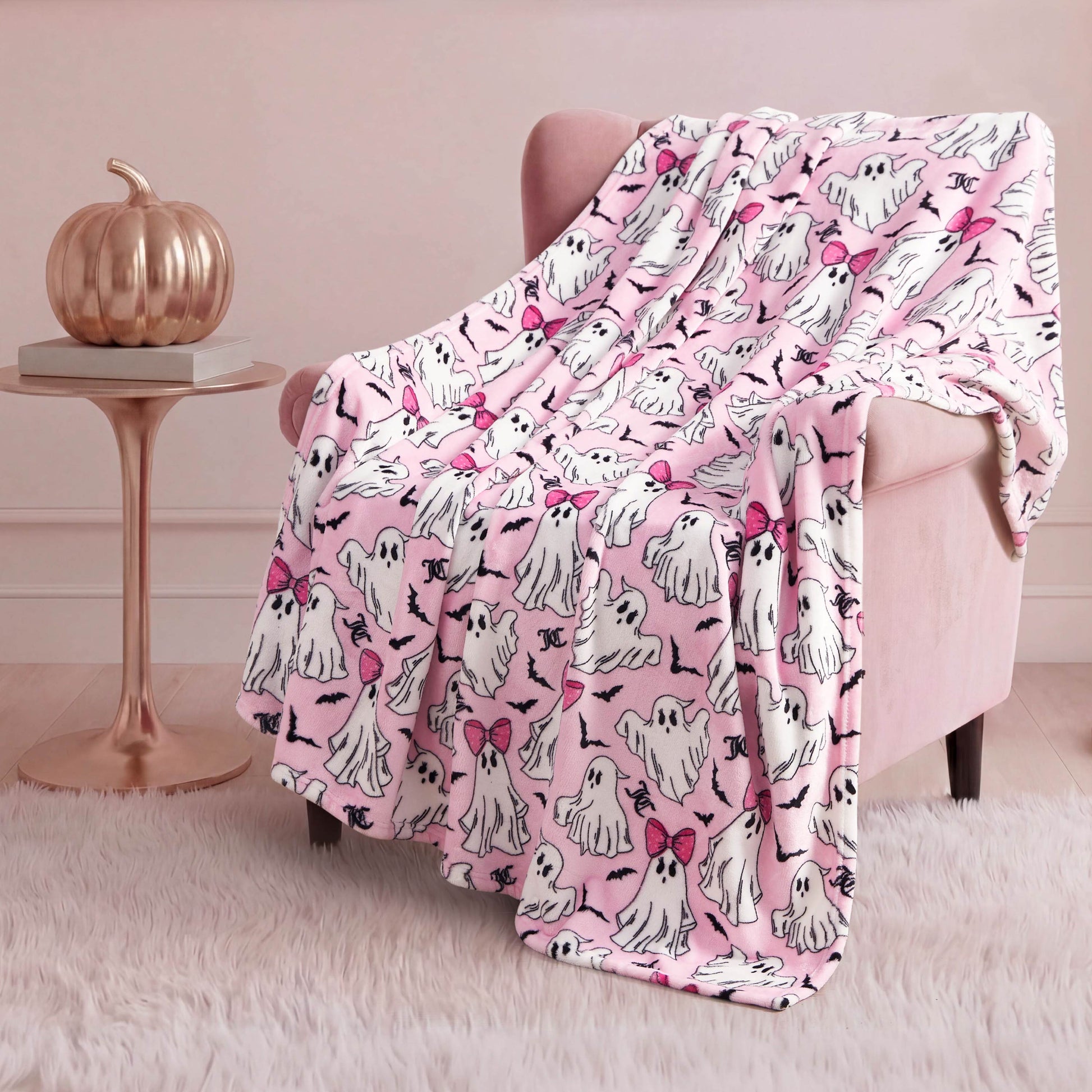 A pink armchair is draped with the Halloween Plush Throw Blanket 50" x 60", featuring cartoon ghosts, some with pink bows. A metallic pumpkin on a round side table and a white rug add a cozy, festive Halloween touch to the room.