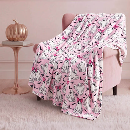 A pink armchair is draped with the Halloween Plush Throw Blanket 50" x 60", featuring cartoon ghosts, some with pink bows. A metallic pumpkin on a round side table and a white rug add a cozy, festive Halloween touch to the room.