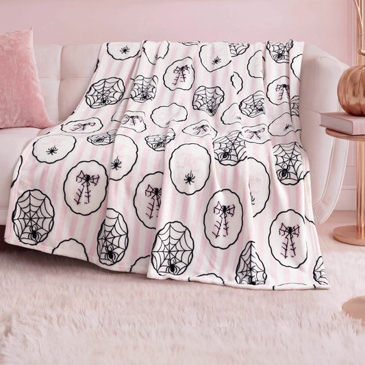 A Halloween Plush Throw Blanket 50" x 60" in white and light pink with black spiderweb, spider, and skeleton bow designs is draped over a light sofa in a chic, pink-themed living room.