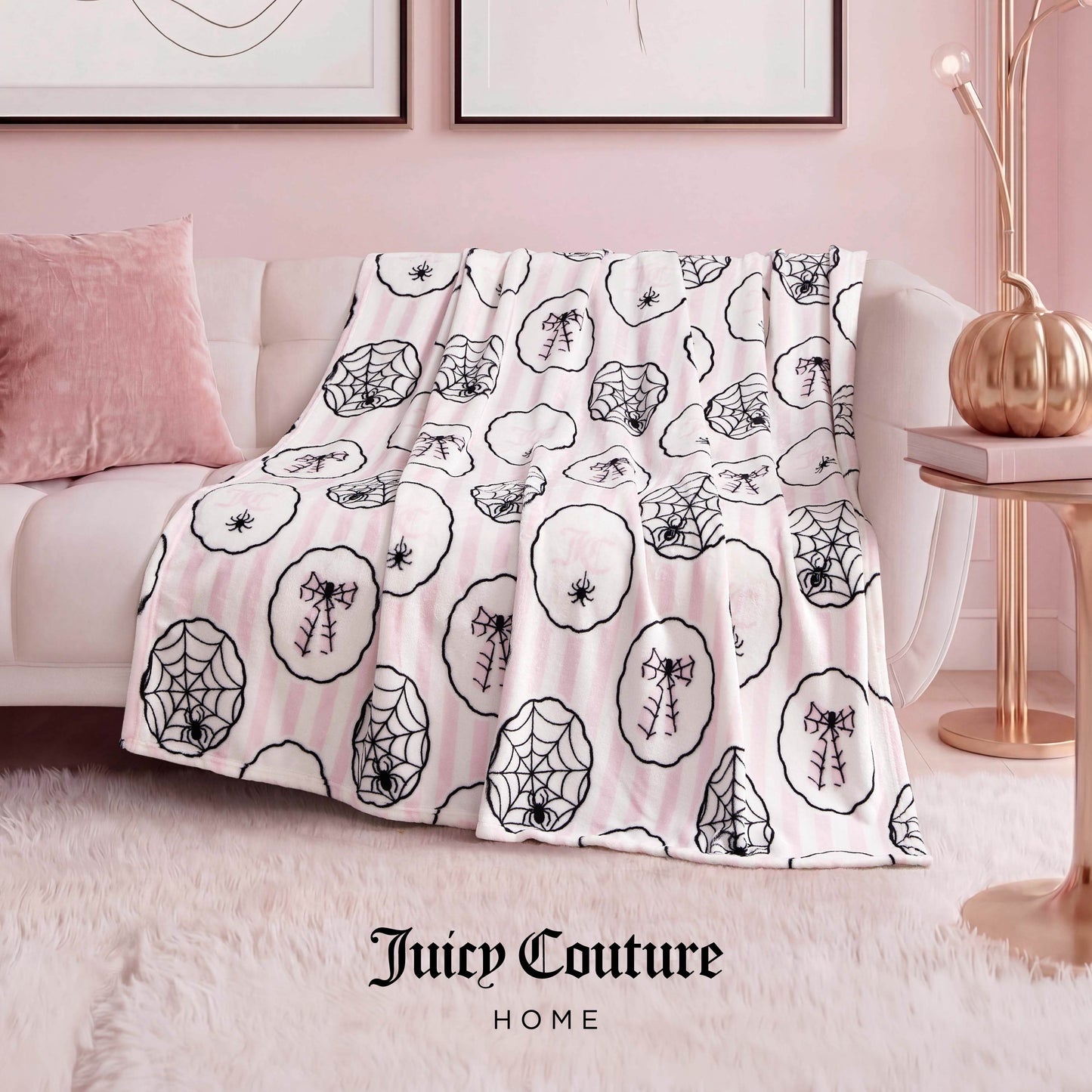The Halloween Plush Throw Blanket 50" x 60" in white and pink stripes, with black spider and web designs, is draped over a white sofa next to a pink pillow in a stylish Juicy Couture Home living room.